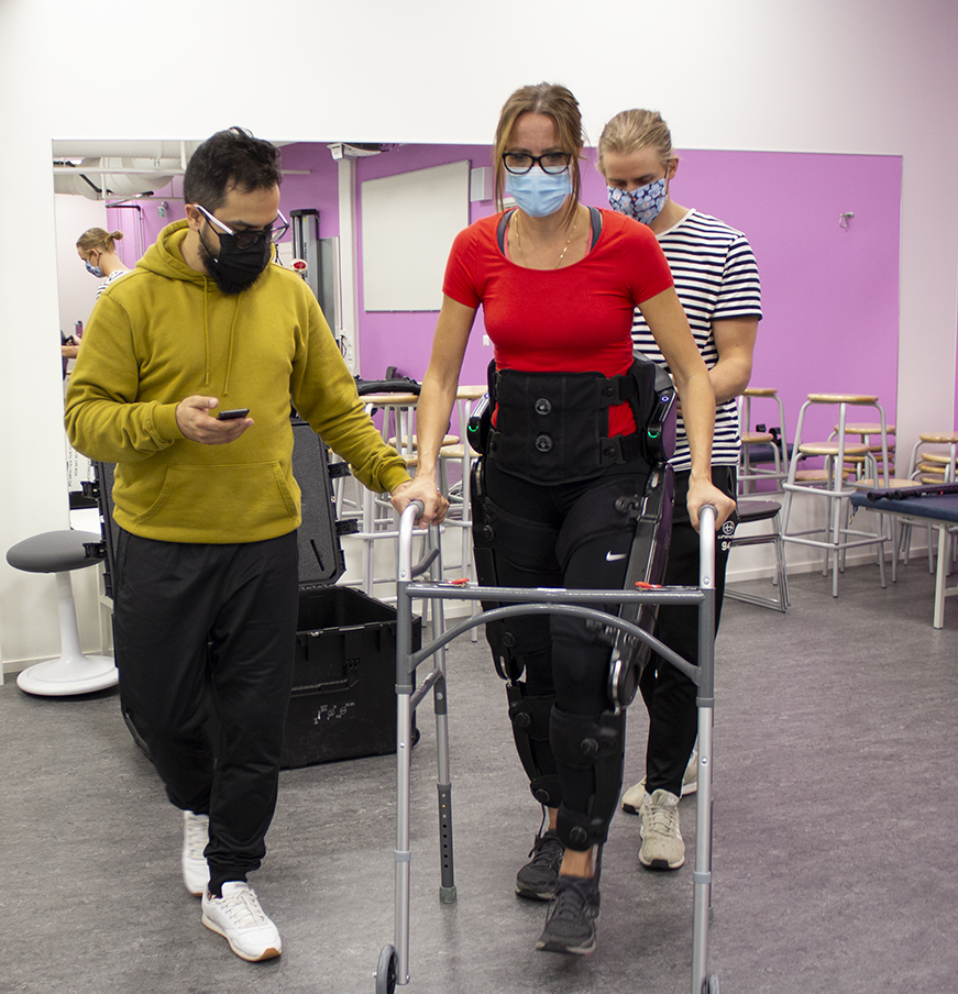 A woman walking with exoskeleton, two men assisting her.
