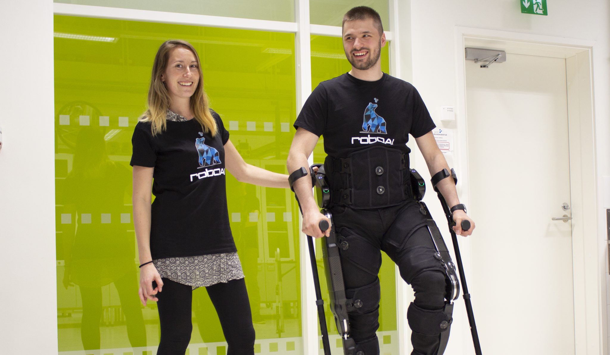 IMG 4738 Juliste The picture shows a woman and a man using an exoskeleton walking robot.