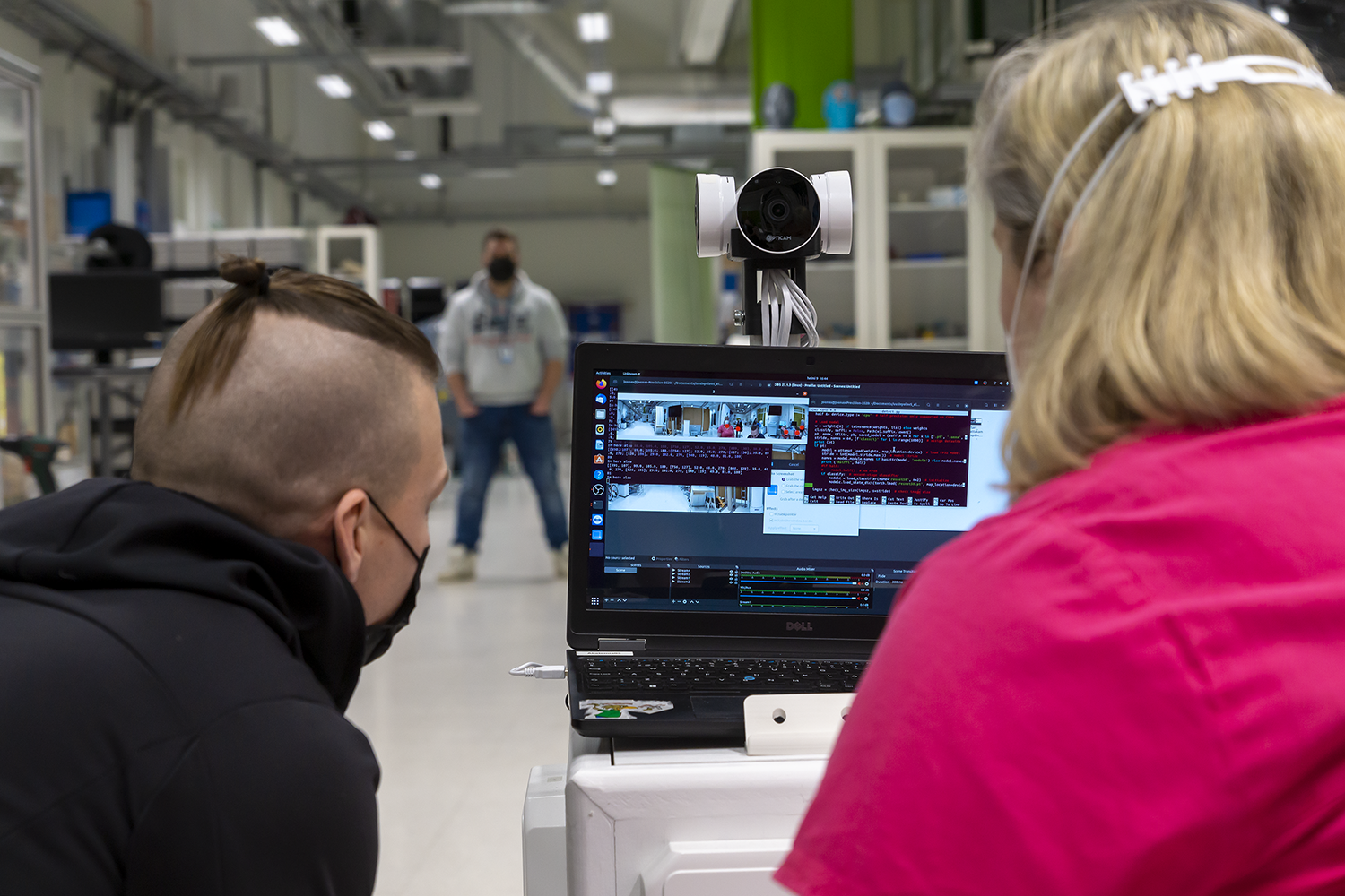 Two people are sitting with their backs to the picture, a computer in front of them and a man standing in the background.