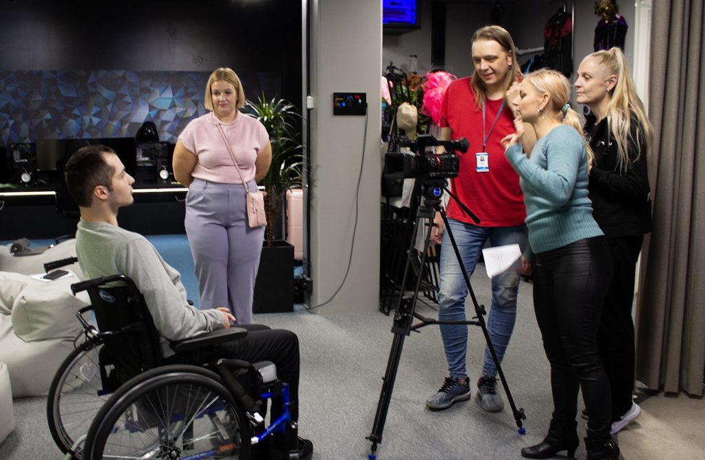 A male person in a wheelchair is in front of the camera. Three people look at what the picture looks like from the camera.