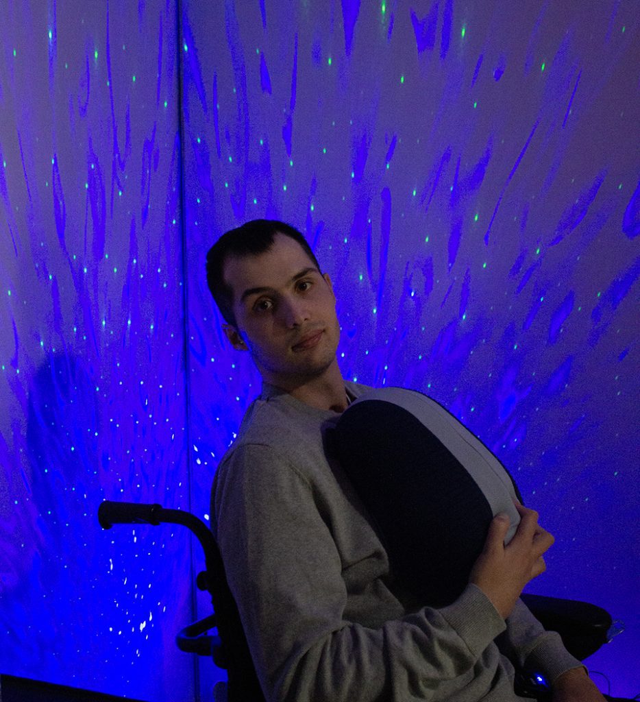 A man is sitting in a wheelchair and holding a sleeping robot in his hands. There is a blue light in the background.