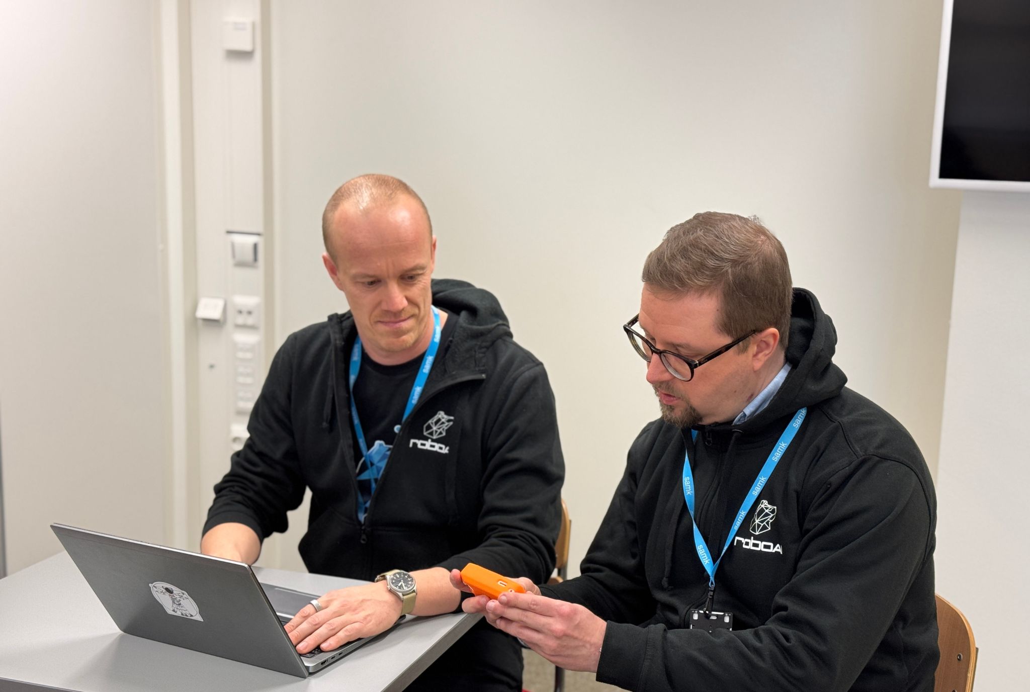 Kyberturvallisuus Kaksi miestä mustissa huppareissa istuu työpöydän äärellä. Vasemman puoleisella miehellä on kannettava tietokone ja oikeanpuoleisella miehellä on laite, jolla voi skannata tietoliikennettä.