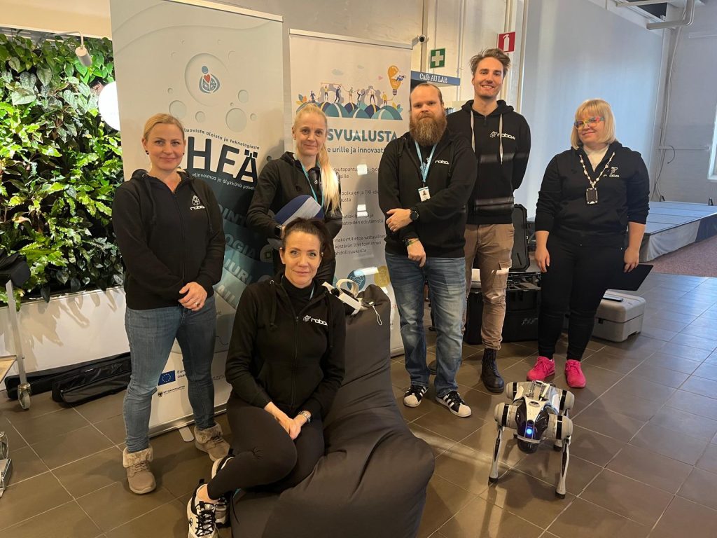 Kuuden henkilön joukko poseeraa kameralle. Vieressä on robottikoira.