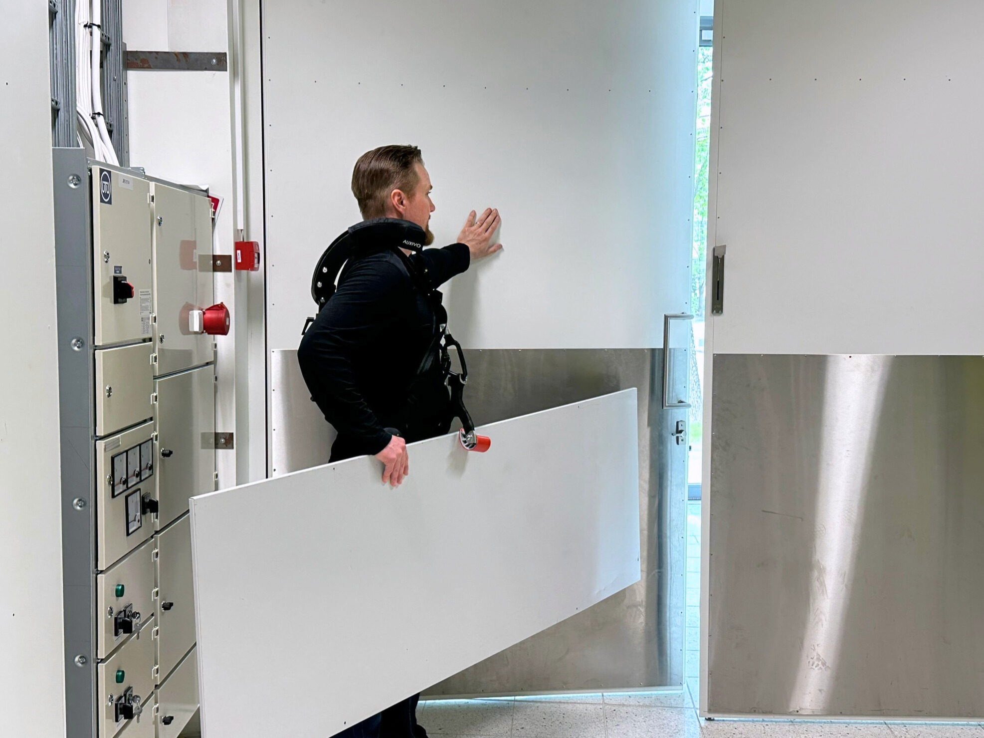 Auxivo carrysuit Mieshenkilö pukeutuneena eksoskeletoniin avaa ovea ja kantaa samalla suurta levyä.