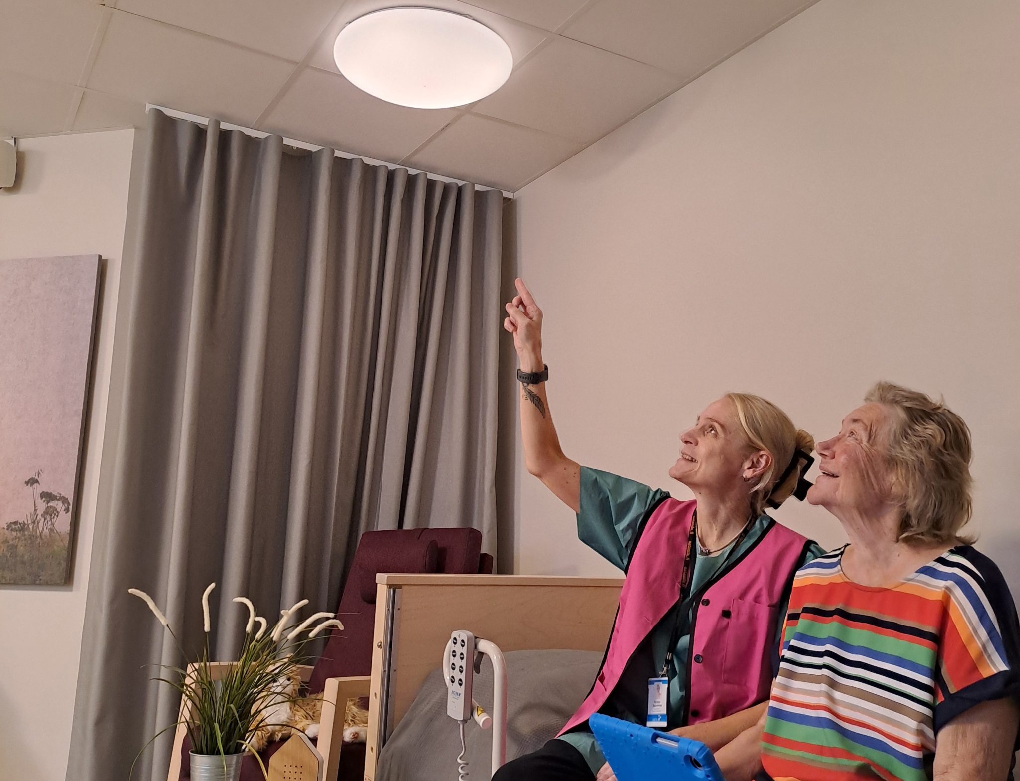Smart Care Muokattu Vaaka Kaksi naista istuu sohvalla ja toinen osoittaa katossa olevaa lamppua.