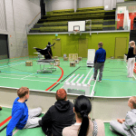 Oppilaita kerääntyneenä seuraamaan kaukalon sisällä tapahtuvaa tehtävää, jossa kolme oppilasta rakentavat esterataa robotille.