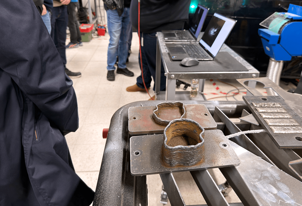 Lähikuva hitsauksen työnjäljestä. Taustalla joukko ihmisiä.