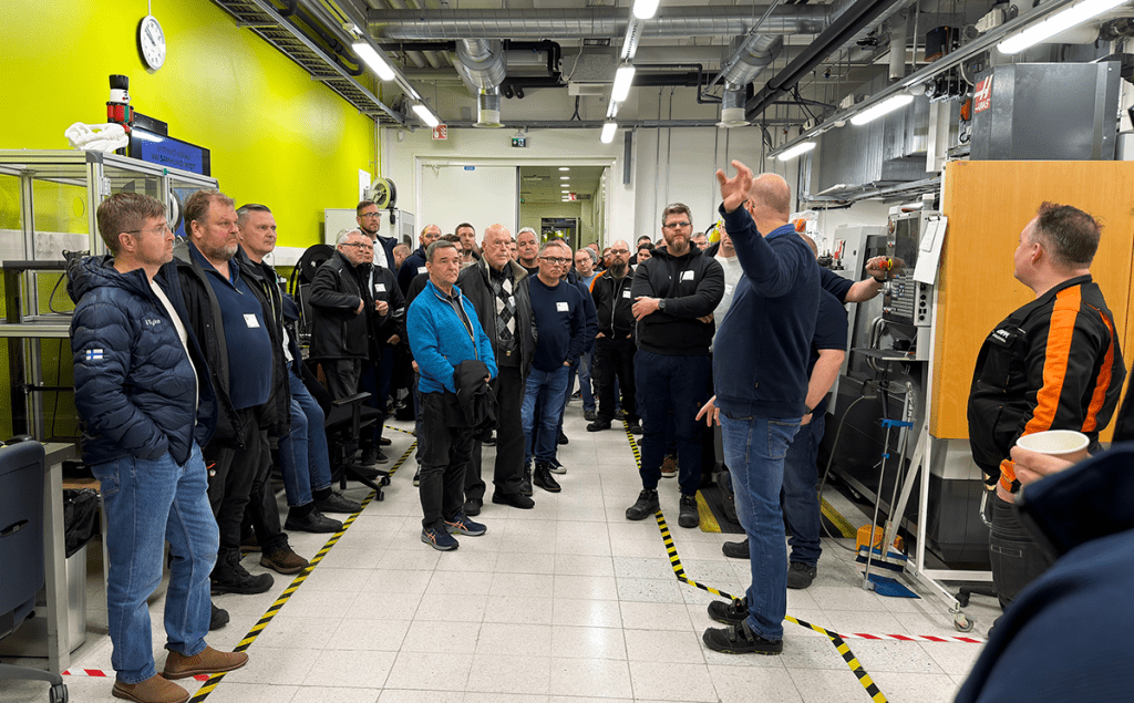 Suuri joukko ihmisiä laboratoriohallissa kuuntelemassa ohjeistusta.