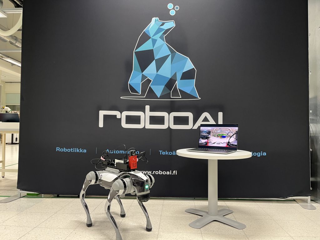 Robottikoira poseeraa kameralle, vieressä kannettavatietokone matalalla pöydällä, jonka näytöllä näkyy videokuvaa.