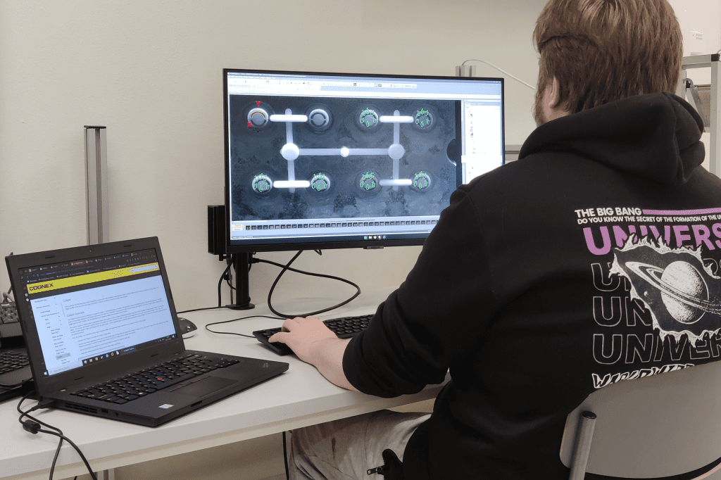 Henkilö istuu kannettavan tietokonen ja näytön äärellä selkä kohti kameraa.