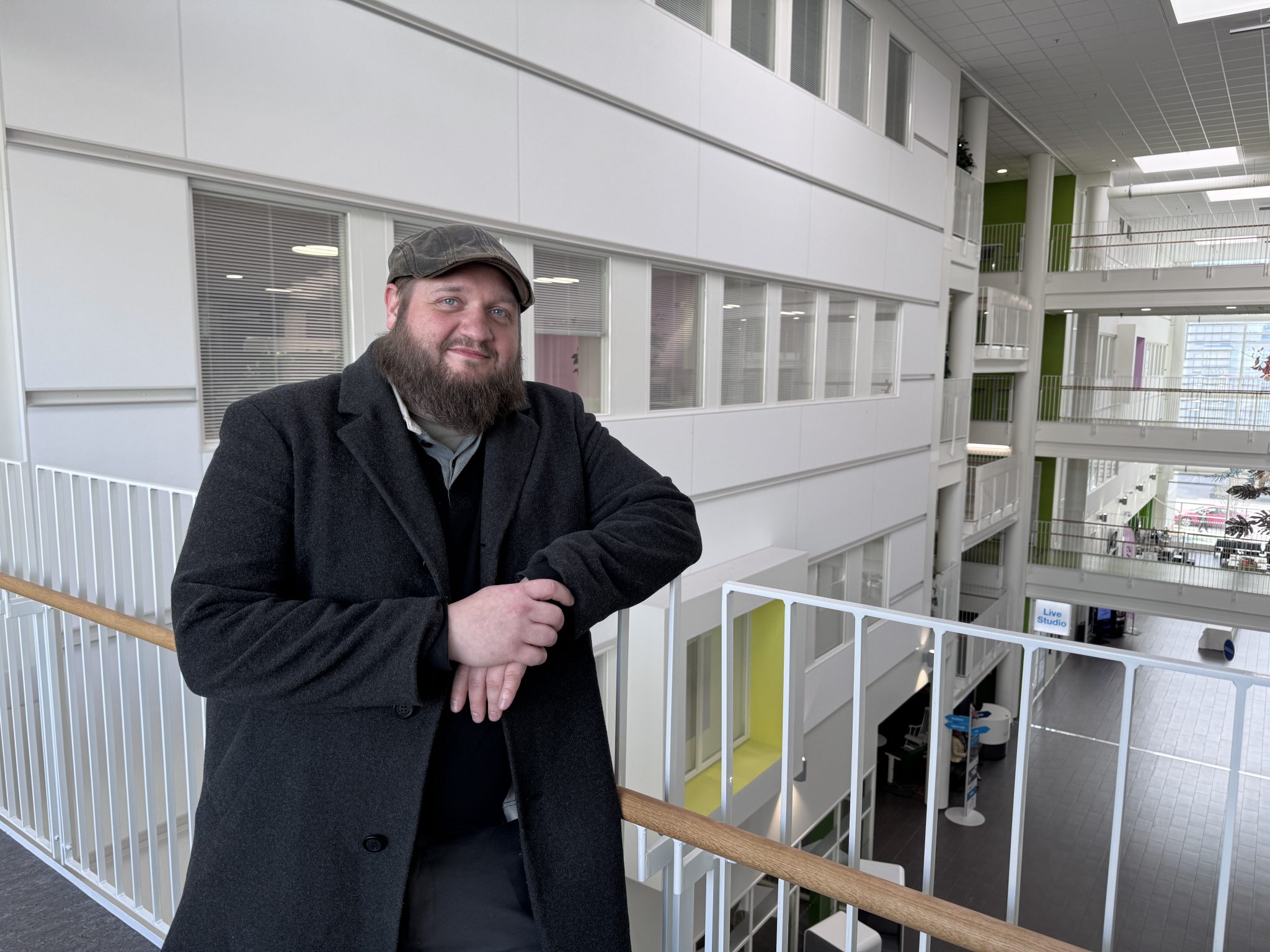 Mies nojaa kaidetta vastaan SAMKissa ja hymyilee. Hänellä on päällään tumma villakangastakki ja päässään lätsä.