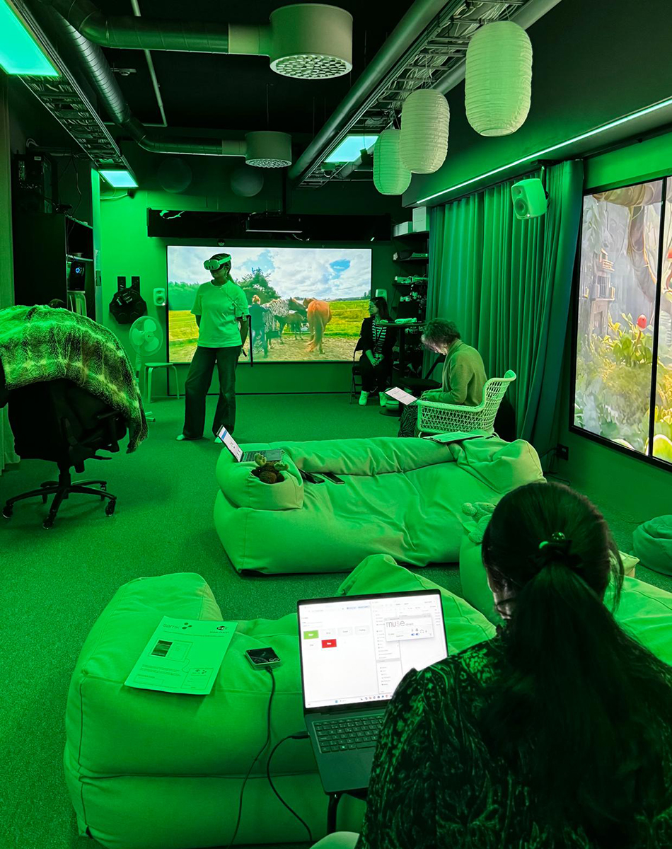 A room lit by green light, where three people are watching the actions of a person wearing VR goggles.