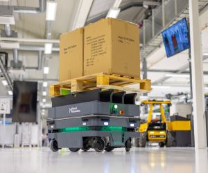 Pahvilaatikkoja kuljettava harmaa nelipyöräinen mobiilirobotti laboratorioympäristössä.