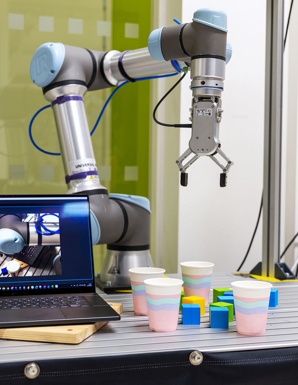 A robot arm with cups and blocks on the table below it, and a laptop next to it.