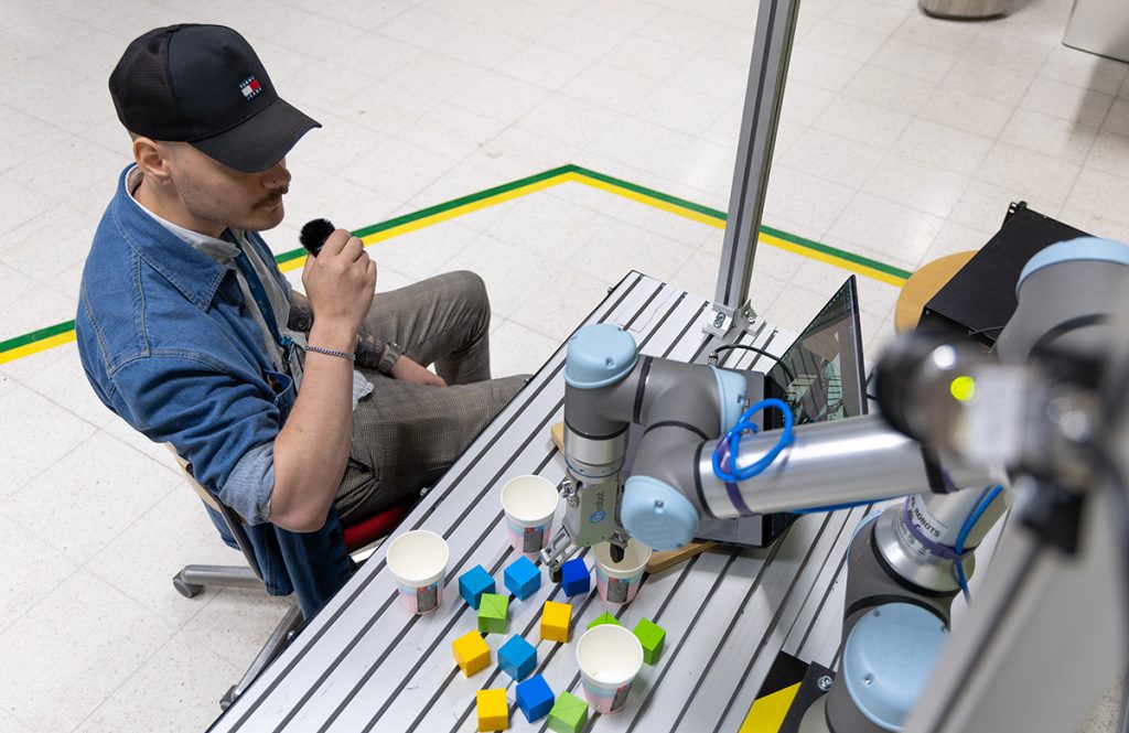 A man is speaking into a microphone in front of a table. On the table are a robot, building blocks, and a laptop.
