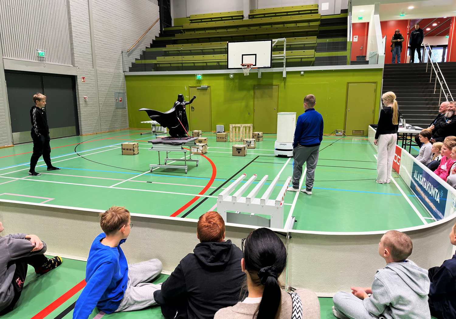 Oppilaita kerääntyneenä seuraamaan kaukalon sisällä tapahtuvaa tehtävää, jossa kolme oppilasta rakentavat esterataa robotille.