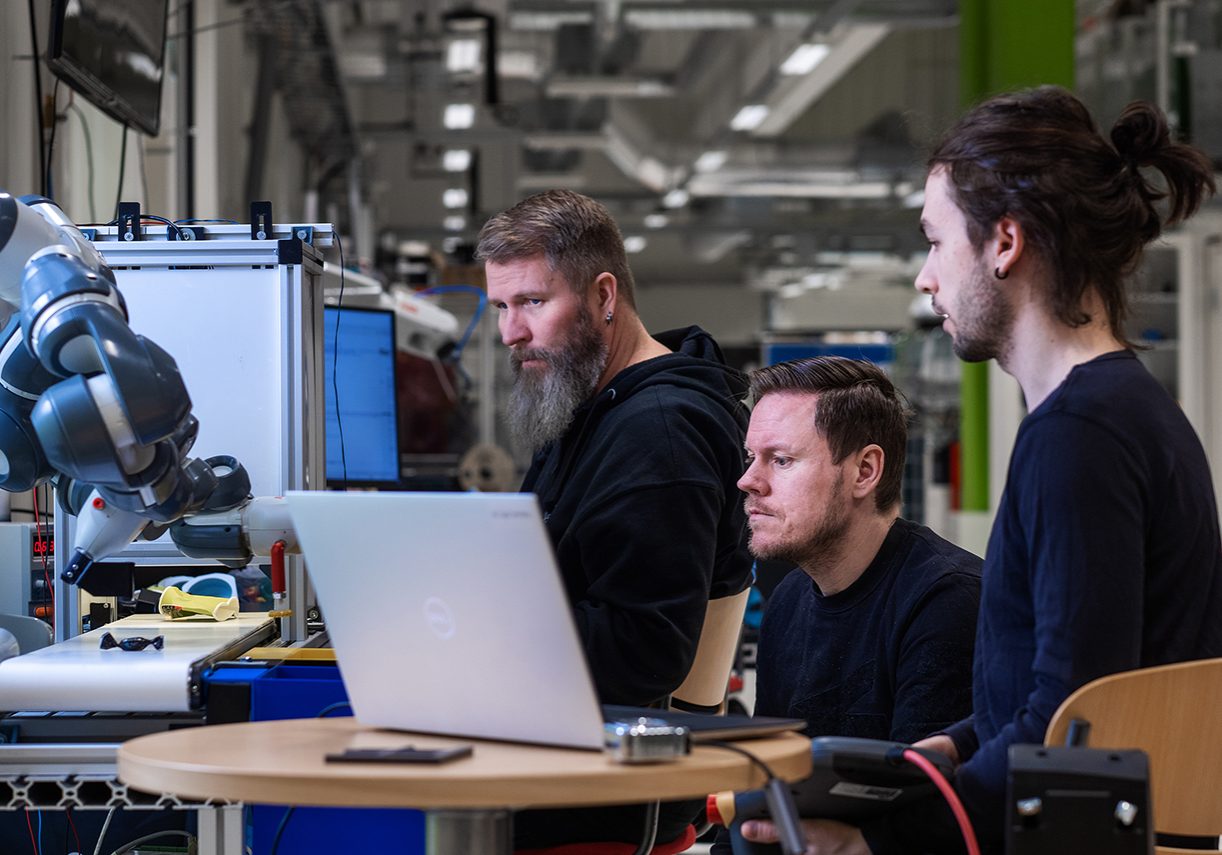 RoboAI Tutkimus Ja Tuotekehityskeskus Kolme akatemialaista työskentelee koneella ja robotilla.