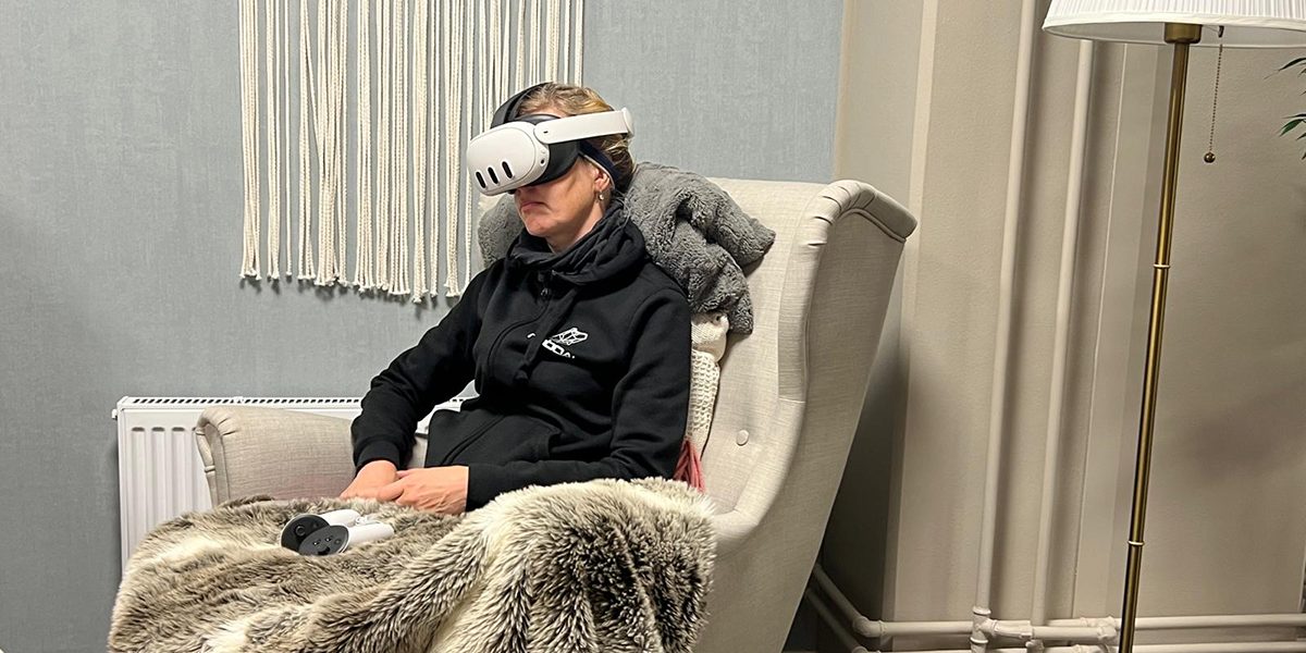 A person is sitting in an armchair, wearing VR goggles and holding a furry blanket.