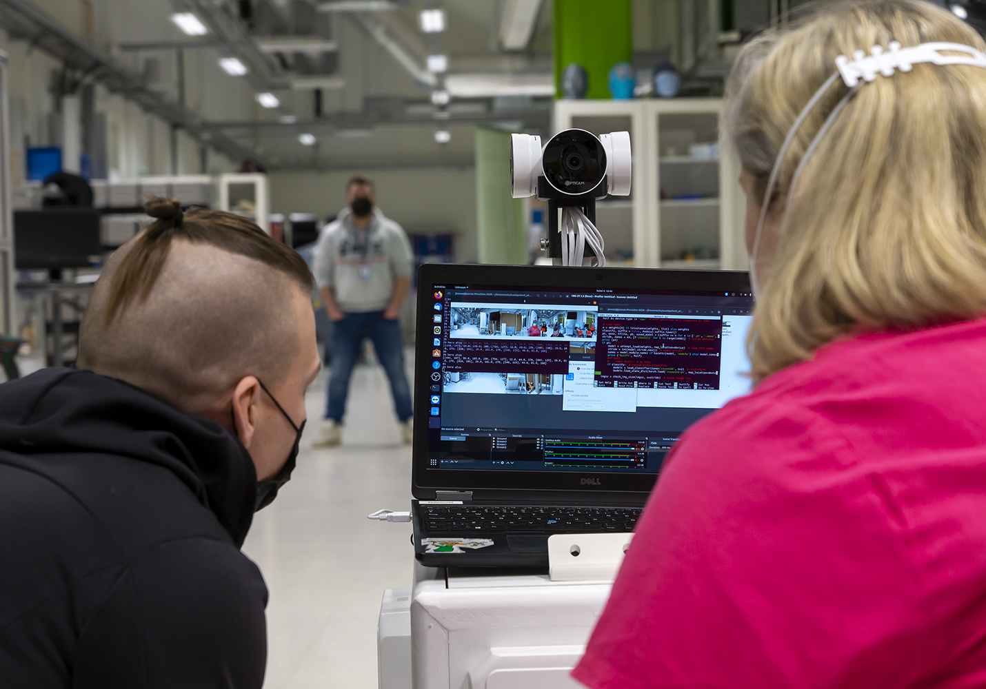 Two people are sitting with their backs to the picture, a computer in front of them and a man standing in the background.